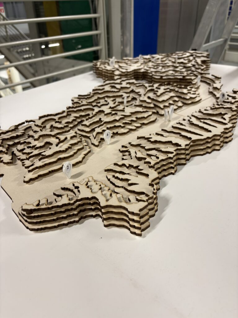 Dokumentacja wystawy „Maintain Mountains – Design for Post-Event Mountain Resilience” na Politecnico di Milano: plansze projektowe „The Vertical Atlas”, model topograficzny z warstwowymi poziomicami oraz przestrzeń ekspozycyjna z dużymi banerami i instalacją w industrialnym wnętrzu kampusu.