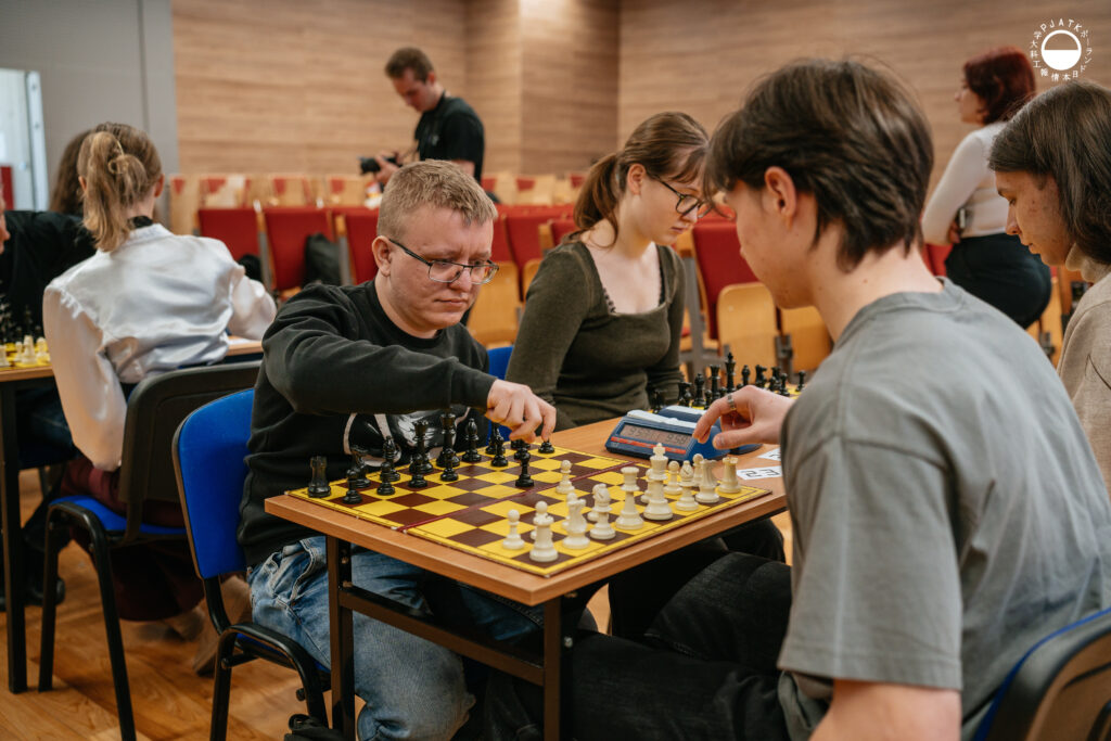 Uczestnicy turnieju grają w szachy przy stolikach w sali.