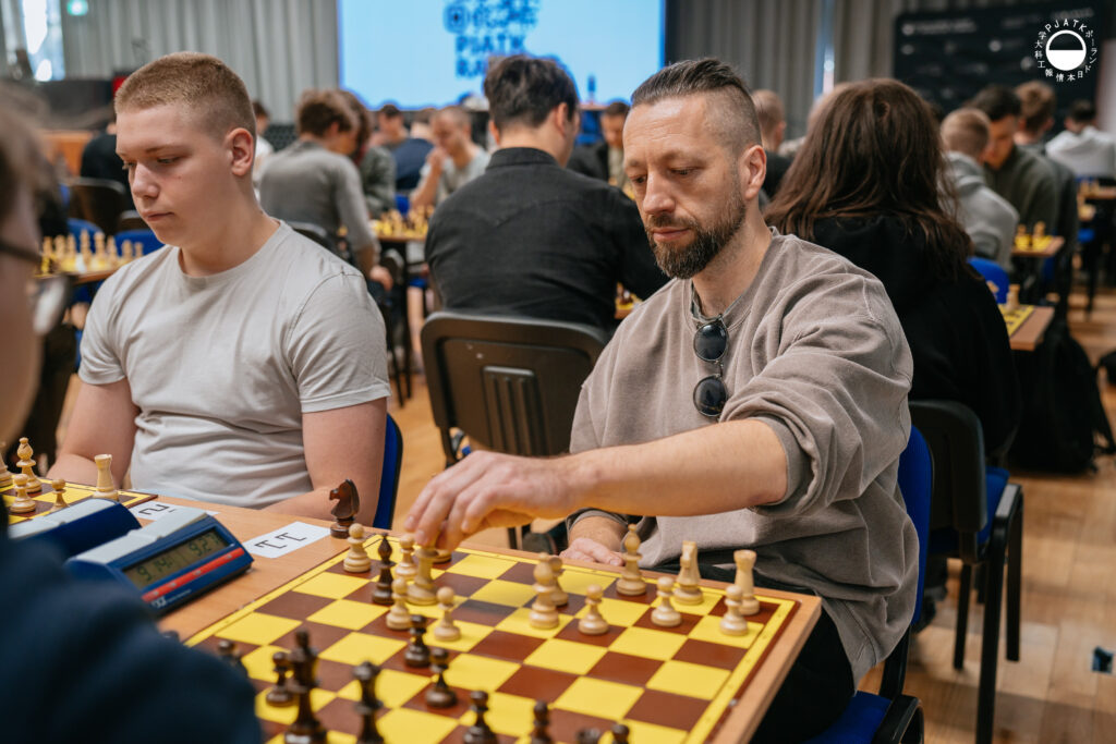 Uczestnicy turnieju grają w szachy przy stolikach w sali.