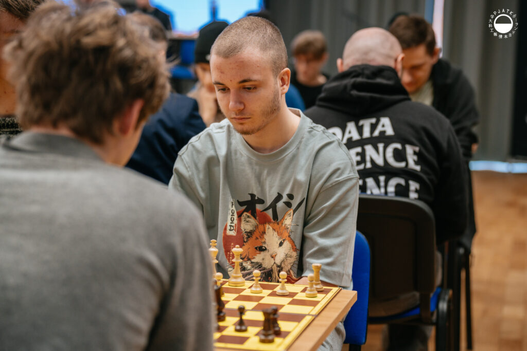 Uczestnicy turnieju grają w szachy przy stolikach w sali.