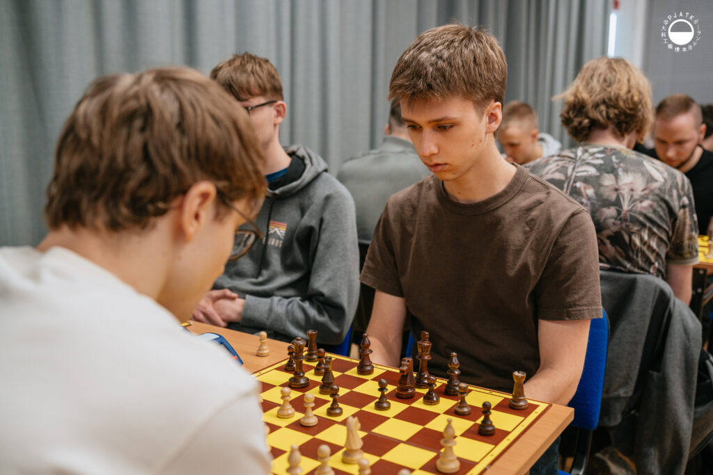 Uczestnicy turnieju grają w szachy przy stolikach w sali.