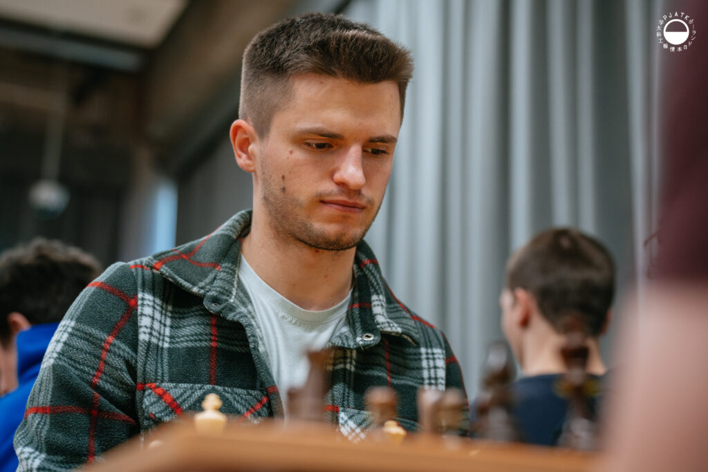 Uczestnicy turnieju grają w szachy przy stolikach w sali.