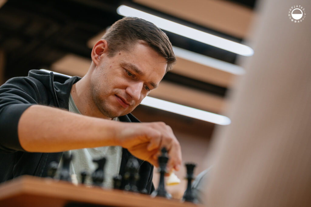 Uczestnicy turnieju grają w szachy przy stolikach w sali.