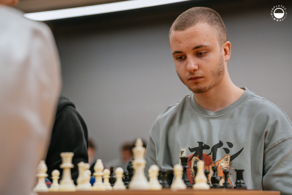 Uczestnicy turnieju grają w szachy przy stolikach w sali.