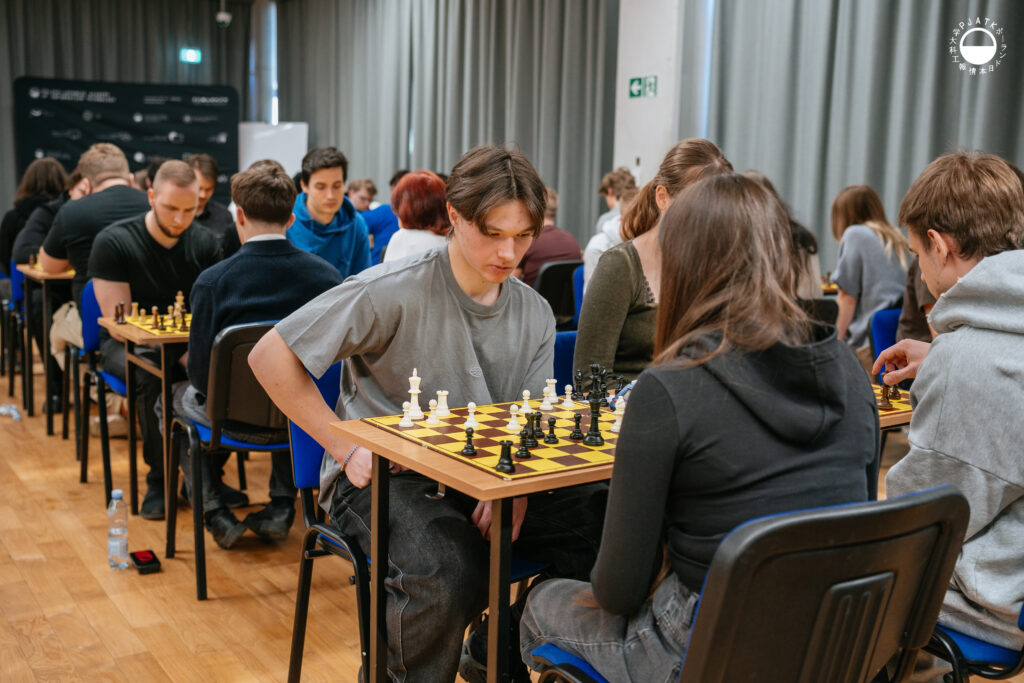 Uczestnicy turnieju grają w szachy przy stolikach w sali.