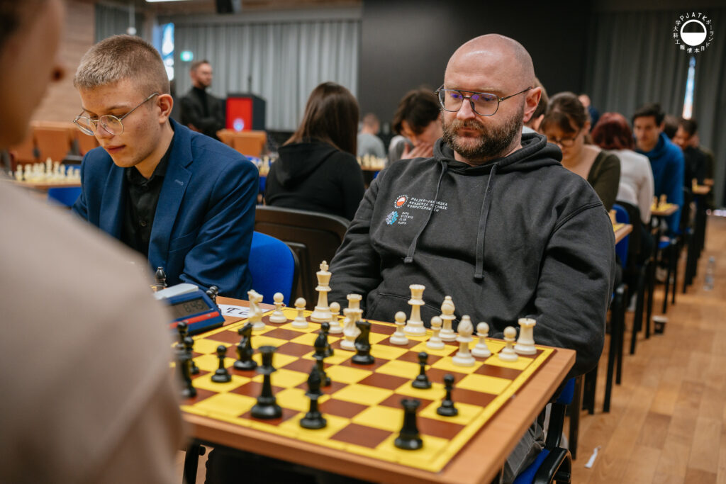 Uczestnicy turnieju grają w szachy przy stolikach w sali.