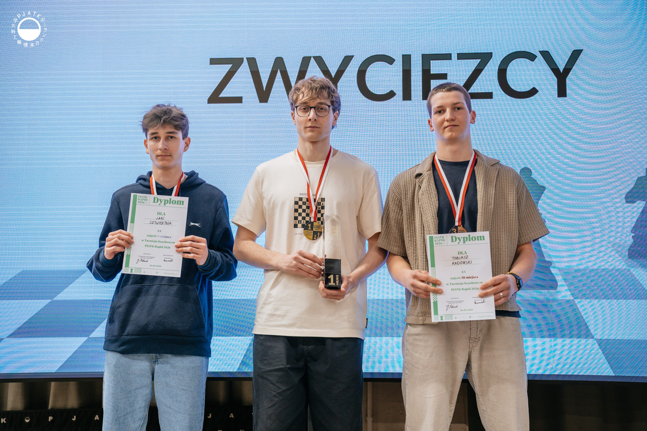 Zwycięzcy turnieju szachowego stoją na scenie z medalami i dyplomami.