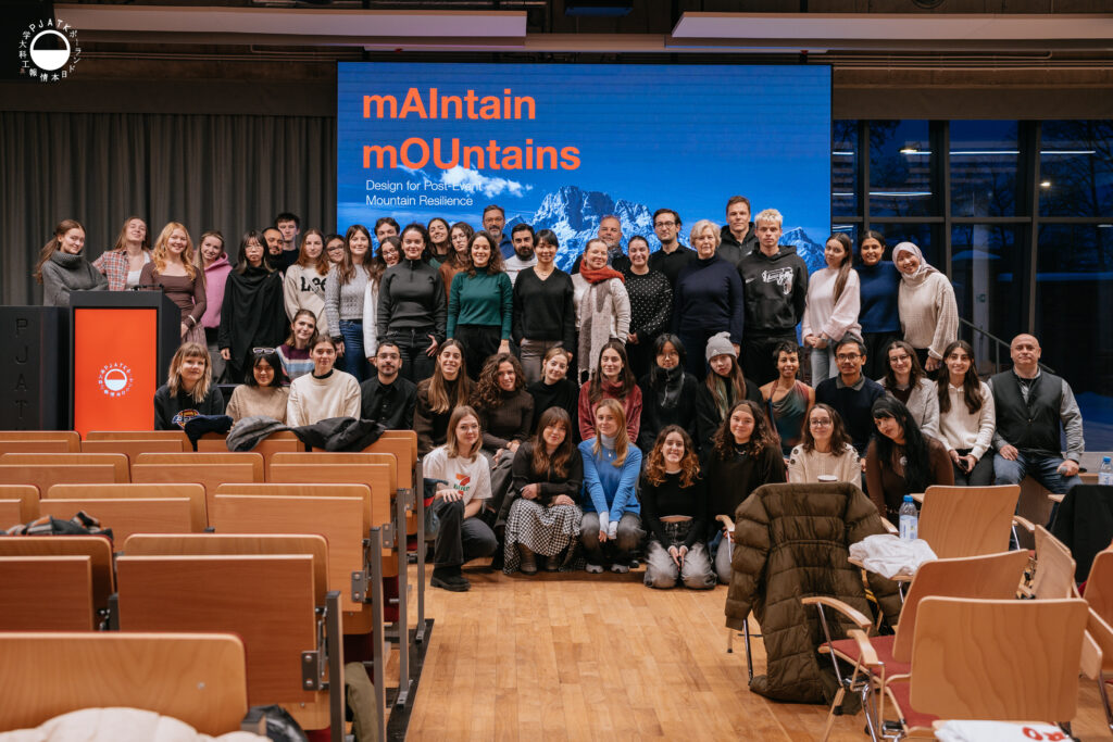 Grupa studentów i wykładowców pozująca do wspólnego zdjęcia w auli Polsko-Japońskiej Akademii Technik Komputerowych przed dużym ekranem z napisem „mAIntain mOUntains – Design for Post-Event Mountain Resilience”.