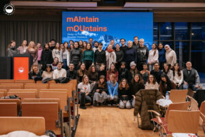 Grupa studentów i wykładowców pozująca do wspólnego zdjęcia w auli Polsko-Japońskiej Akademii Technik Komputerowych przed dużym ekranem z napisem „mAIntain mOUntains – Design for Post-Event Mountain Resilience”.