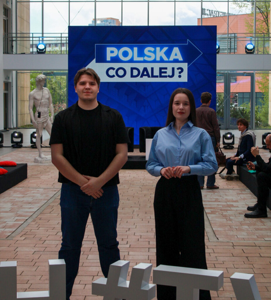 (dwie osoby w holu z napisem) Studenci stoją w przestrzeni wydarzenia przed sceną z napisem „Polska. Co dalej?” przygotowaną do debaty.