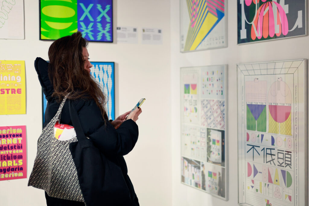 Kobieta fotografuje lub ogląda plakaty typograficzne prezentowane na ścianie galerii.