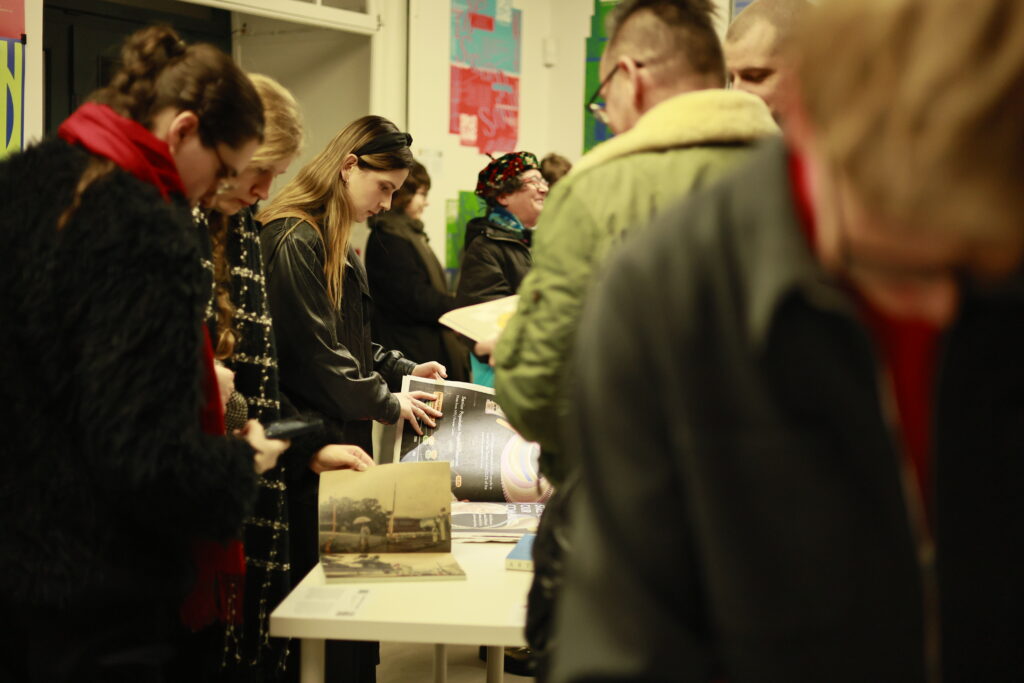 Grupa osób ogląda i przegląda projekty typograficzne wyłożone na stole podczas wystawy.