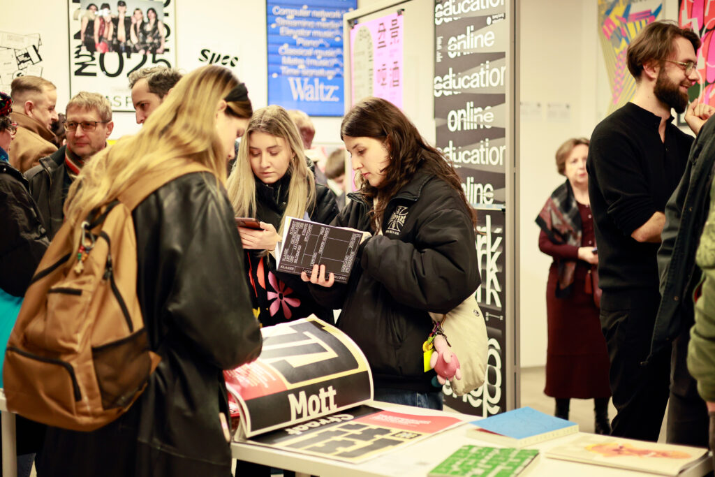 Grupa osób ogląda publikacje i rozmawia przy stoisku z projektami typograficznymi.