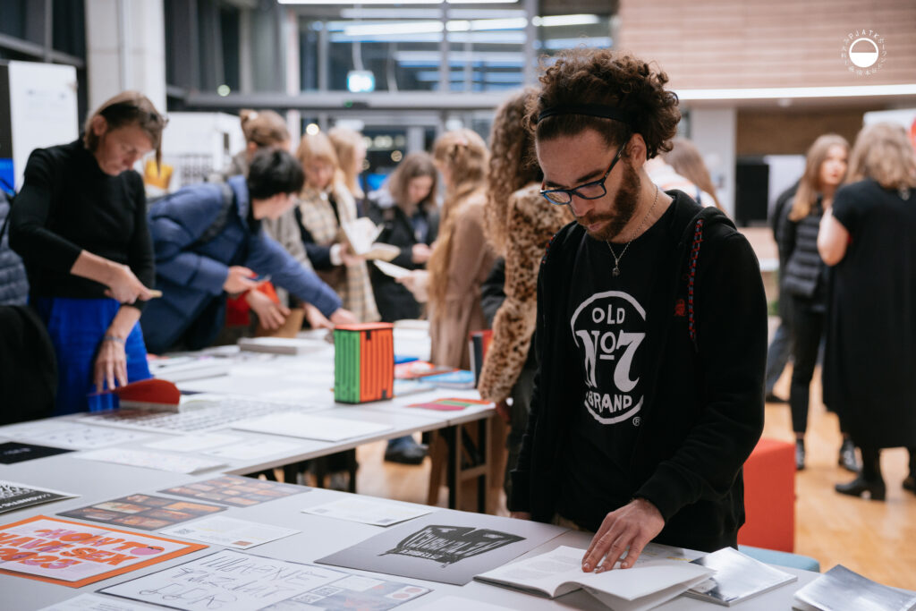 Mężczyzna przegląda publikację typograficzną przy stole wystawowym wśród innych uczestników.