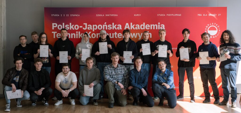A group of students stands in front of the wall at the Polish-Japanese Academy of Information Technology, holding their cybersecurity diplomas.