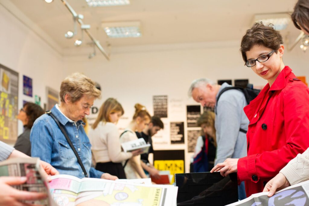 Kobieta w czerwonym płaszczu przegląda publikację wśród innych uczestników wystawy.