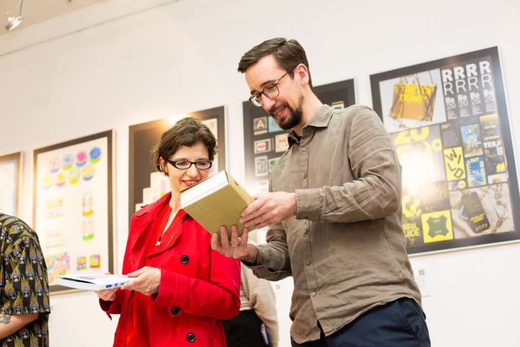 Dwie osoby oglądają książkę i rozmawiają na tle wystawy projektów typograficznych.
