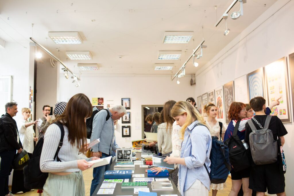 Uczestnicy wystawy przeglądają książki i projekty typograficzne wyłożone na stołach w galerii.