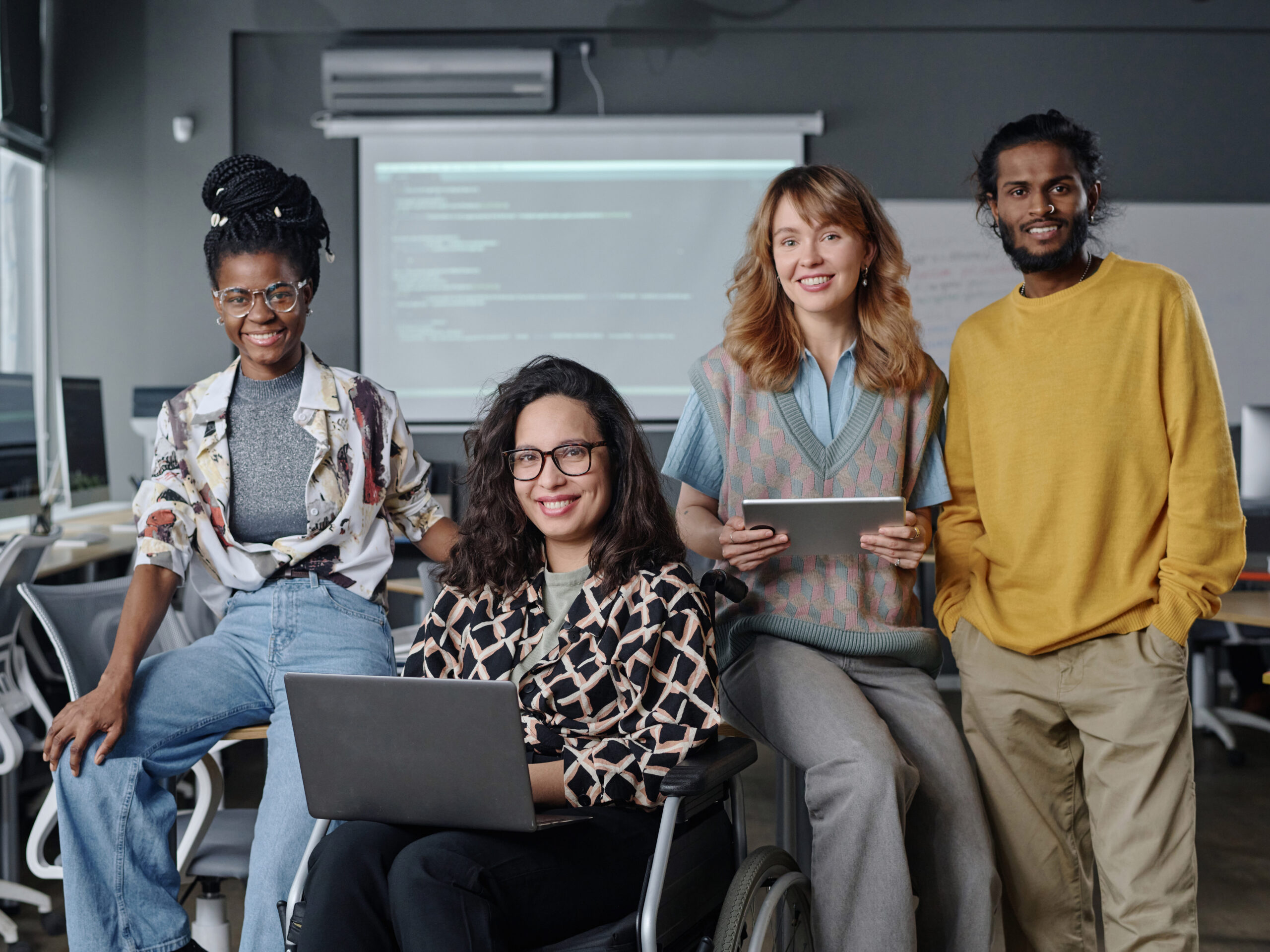 Diverse group of young programmers posing together at office holding gadgets and smiling at camera