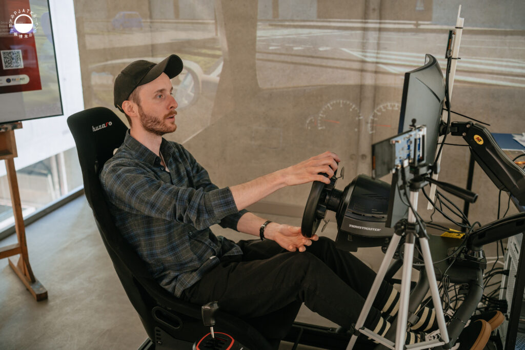 Zdjęcia z wydarzenia „Tokyo vibe, Warsaw drive” w PJATK. Uczestnicy biorą udział w wykładach oraz testują symulator jazdy samochodem przygotowany w ramach projektu Katedry Multimediów. Na fotografiach widać osoby korzystające z kierownicy i ekranów symulatora, widok wirtualnej trasy, prelegentów występujących w auli oraz uczestników spotkania.