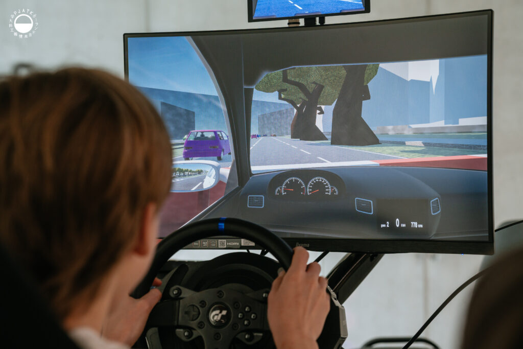 Zdjęcia z wydarzenia „Tokyo vibe, Warsaw drive” w PJATK. Uczestnicy biorą udział w wykładach oraz testują symulator jazdy samochodem przygotowany w ramach projektu Katedry Multimediów. Na fotografiach widać osoby korzystające z kierownicy i ekranów symulatora, widok wirtualnej trasy, prelegentów występujących w auli oraz uczestników spotkania.