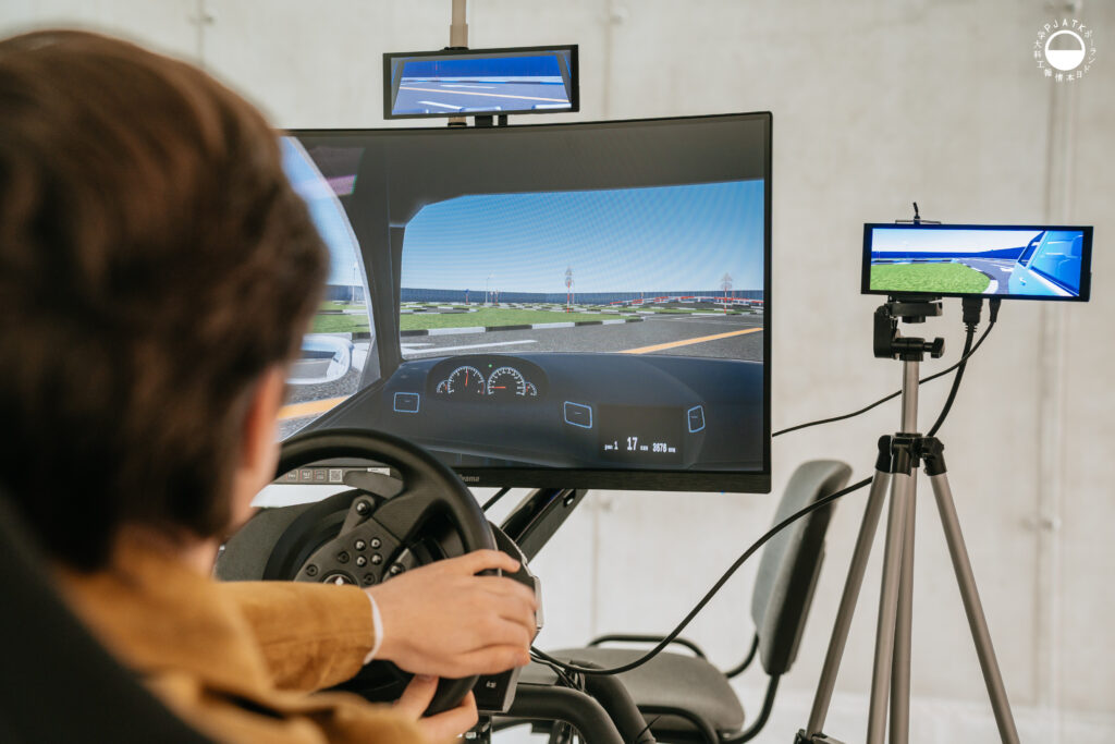 Zdjęcia z wydarzenia „Tokyo vibe, Warsaw drive” w PJATK. Uczestnicy biorą udział w wykładach oraz testują symulator jazdy samochodem przygotowany w ramach projektu Katedry Multimediów. Na fotografiach widać osoby korzystające z kierownicy i ekranów symulatora, widok wirtualnej trasy, prelegentów występujących w auli oraz uczestników spotkania.