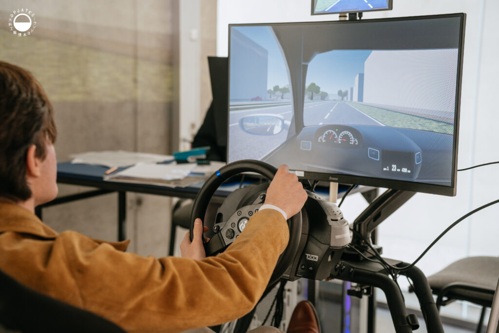 Zdjęcia z wydarzenia „Tokyo vibe, Warsaw drive” w PJATK. Uczestnicy biorą udział w wykładach oraz testują symulator jazdy samochodem przygotowany w ramach projektu Katedry Multimediów. Na fotografiach widać osoby korzystające z kierownicy i ekranów symulatora, widok wirtualnej trasy, prelegentów występujących w auli oraz uczestników spotkania.