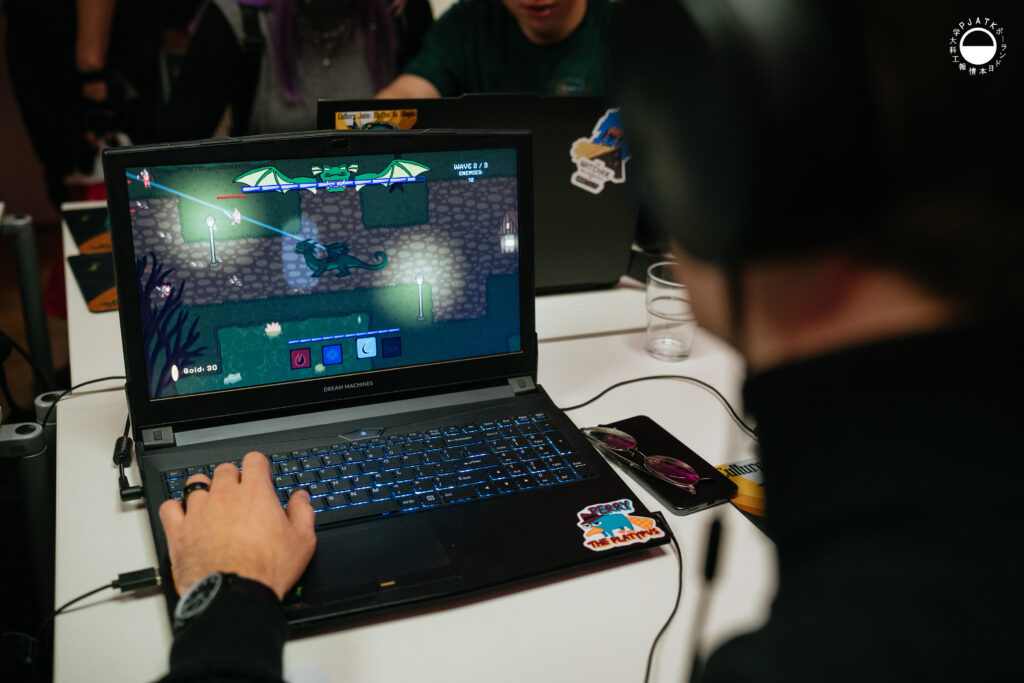 Osoba grająca na laptopie podczas wydarzenia, na ekranie widoczna kolorowa gra komputerowa, a w tle inni uczestnicy siedzący przy komputerach.