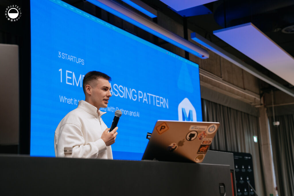 Prelegent z mikrofonem prowadzi prezentację przy mównicy w auli PJATK. W tle widoczny jest slajd dotyczący startupu i przetwarzania danych z użyciem Pythona oraz AI.