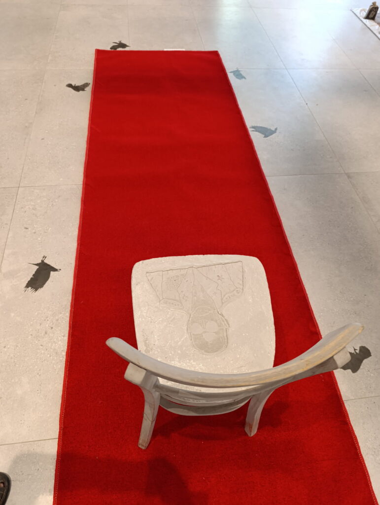 Puste krzesło stoi na czerwonym dywanie w przestrzeni galerii, otoczone sylwetkami czarnych ptaków na podłodze.