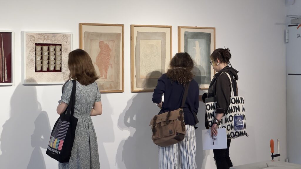 Zwiedzający oglądają cykl grafik Andrzeja Kaliny prezentowanych w drewnianych ramach na ścianie galerii.