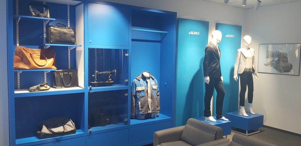 Showroom firmy JUKI z ekspozycją ubrań, toreb i maszyn do szycia. W tle widoczne są niebieskie ściany wystawiennicze z logo JUKI oraz manekiny prezentujące odzież.