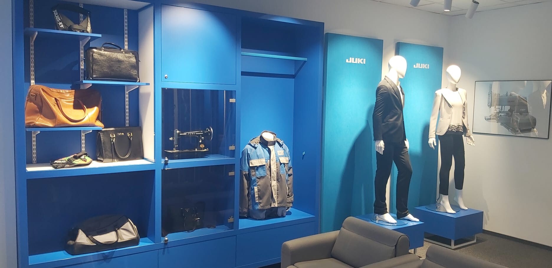 Showroom firmy JUKI z ekspozycją ubrań, toreb i maszyn do szycia. W tle widoczne są niebieskie ściany wystawiennicze z logo JUKI oraz manekiny prezentujące odzież.